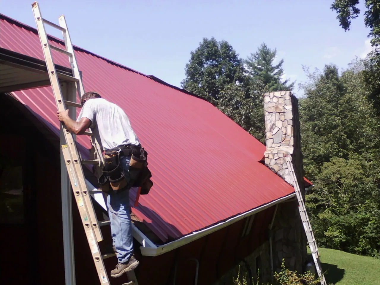 Roofer on a ladder beside a red metal roof and stone chimney — Soffit & Fascia Repair by Roofing Brothers in East Waterboro